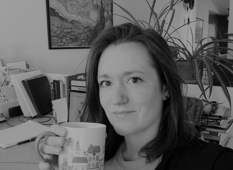 Black and white photo of Nicole Patrie. A light skinned woman with dark hair, looking at the camera with a slight smile and holding a coffee mug. In the background are books, plants, and a photo on the wall.
